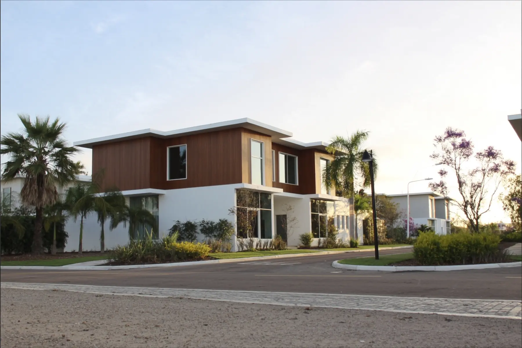 Casa moderna de dos plantas en blanco y madera, en una calle residencial arbolada al atardecer