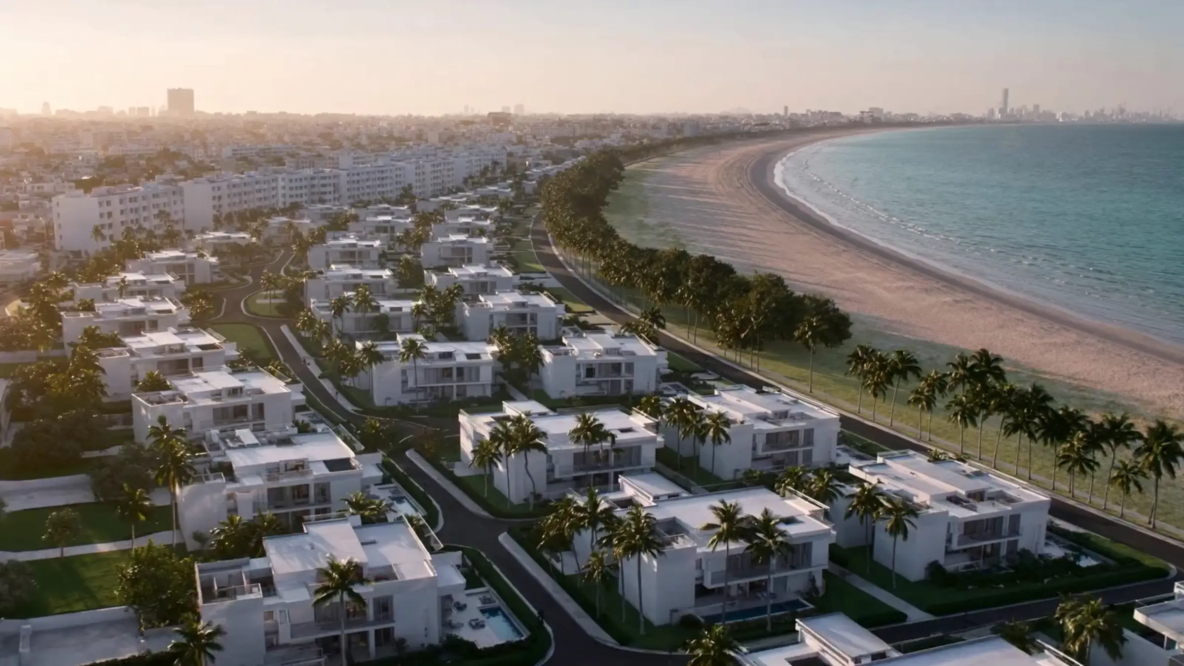 Vista aérea de un desarrollo residencial costero al atardecer, con villas blancas, palmeras y el mar al fondo