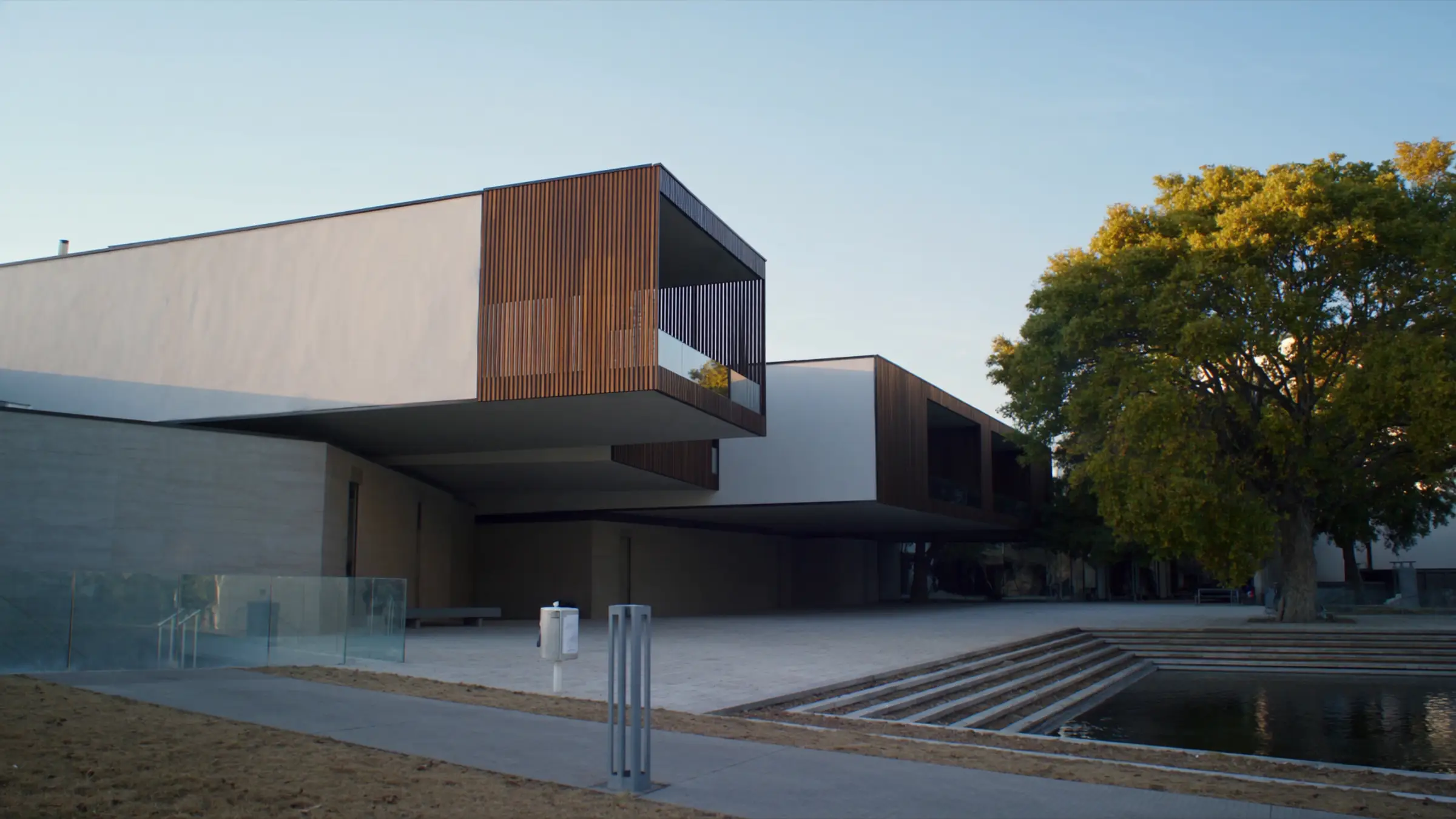 Edificio moderno de baja altura en blanco y madera, con una plaza ajardinada y un espejo de agua al atardecer